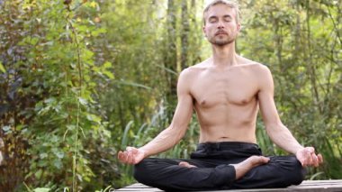 Genç adam pratik yoga açık sakallı. Onun oturma poz lotus ile gözleri kapalı ve meditasyon. O zaman birlikte ellerini kıvrımlar ve şükran olarak yay. Zemin üzerine yeşil orman.