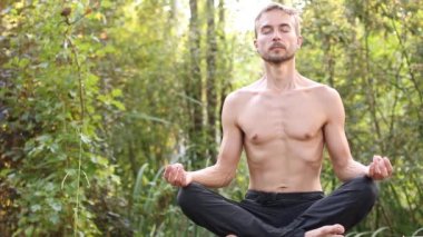 Lotus poz gözleri kapalı oturan genç adam. O nefes derin, sonra gülümseyerek gözlerini açmak ve etrafına bak. Yeşil orman arka plan üzerinde. Yoga doğa kavramı.