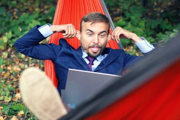 Businessman with laptop relaxs in a hammock on nature. He talking on video chat and doing funny faces with his tongue hanging out and splayed ears. Freelance or telework, on-line chatting concept.