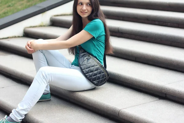 Long haired millenial woman in mint t-shirt and white jeans with modern leather bag sitting on stairs. Candid lifestyle photo. College or city life concept.