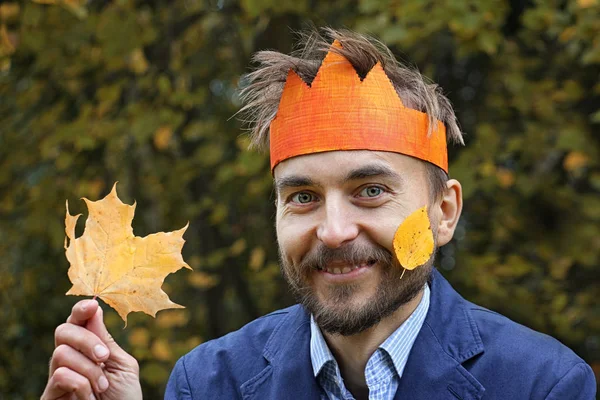 King of the fall. Funny bearded man in paper crown with yellow leaf on his cheek and in his arm. Autumn forest on a background. Seasonal concept.