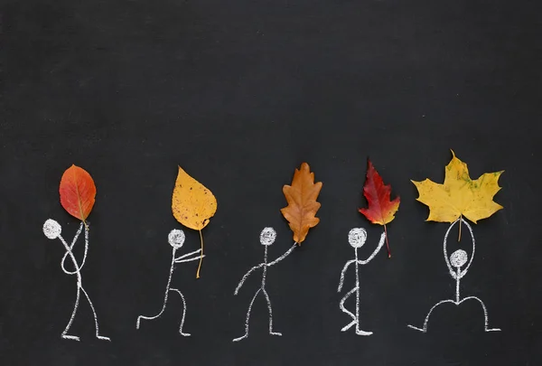 Five people, drawn in chalk, they are in different poses and hold real autumn leaves in their hands, on blackboard or chalkboard background. Fall concept. Copy space. Good for postcard.
