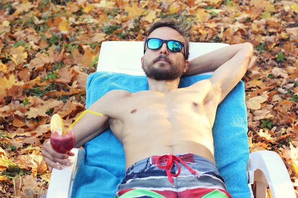 Man in swimming shorts and sunglasses, with cocktail is lying on a sun lounger in the middle of a lawn covered with fallen leaves. Fall vacation or holiday in the autumn. Seasonal travel concept.
