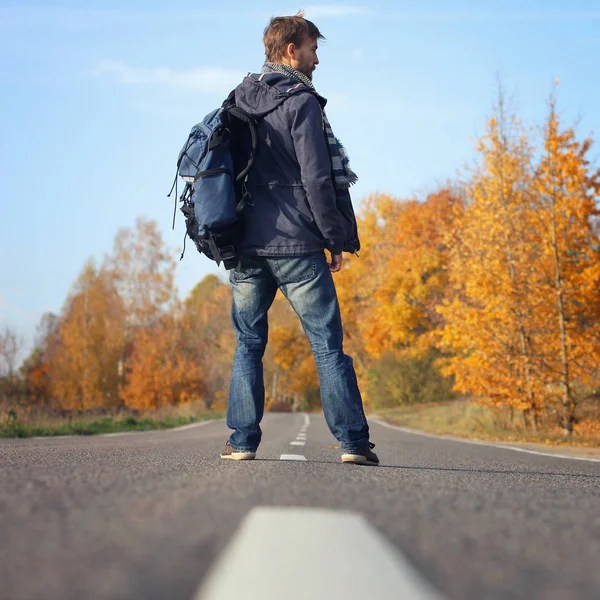 Handsome hipster anventurer in casual wear with backpack walking on the empty road, surrounded by trees with yellow leaves. Discovery beautiful fall season. Autumn travel concept. Square.