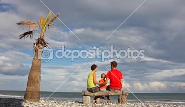 Aile tatil. Anne, baba ve onun çocuğunu yalnız palmiye ağacının altında sahilde ahşap bankta sarılma ve birbirlerini öpmeye oturuyor.