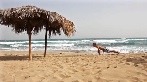 年轻的帅哥在海滩上做俯卧撑 靠近大甘蔗太阳伞 健身中心 — 图库视频影像