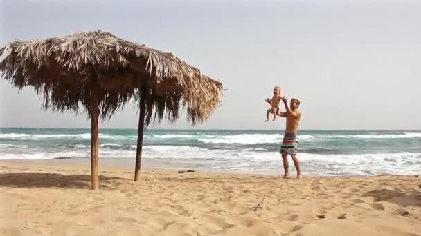 父子上下座在海滩上玩得很开心 附近有大的藤条太阳伞 爸爸用手抱着孩子把他扔了起来 为人父母和家庭旅行概念 — 图库视频影像