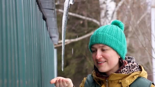 微笑的女孩戴着绿色针织帽子 看着一个大的融化的冰柱 把她的手放在水滴下 春天的概念 — 图库视频影像