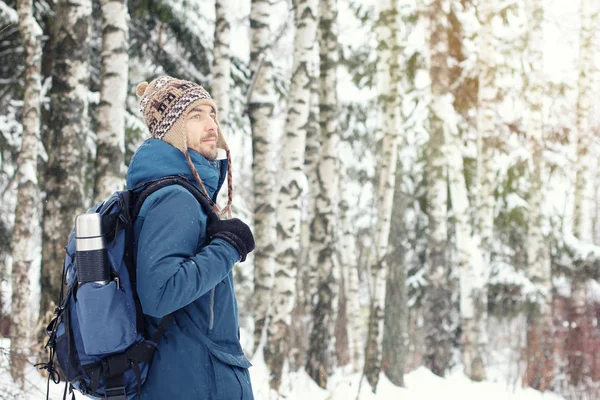 バックパックと魔法瓶と暖かい服でハンサムなひげを生やした男性ハイカーは 雪に覆われた冬の森で旅行します 季節の屋外の旅行の概念 コピー スペース — ストック写真