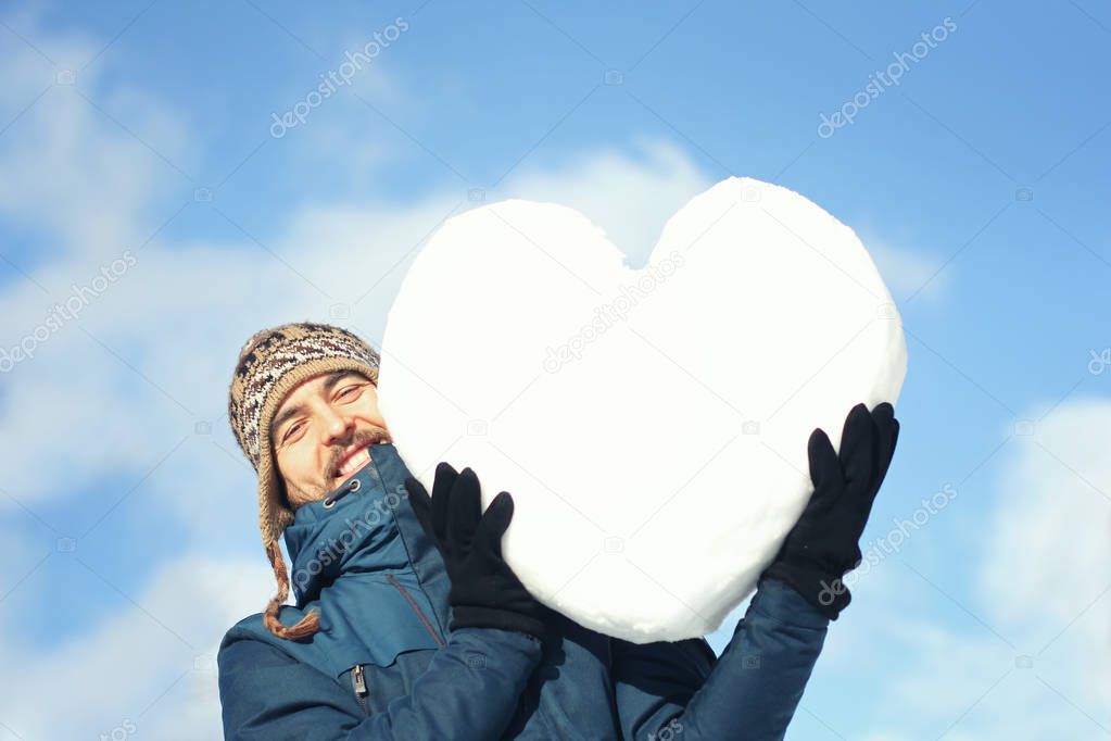 Handsome smiling man in winter clothes holding a big heart made of snow, on the blue sky background. Declarations of love, valentines, international womens or good deeds day concept. 