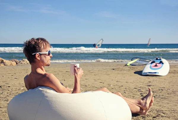 Мужчина в солнечных очках отдыхает на пуфе на пляже с чашкой t — стоковое фото