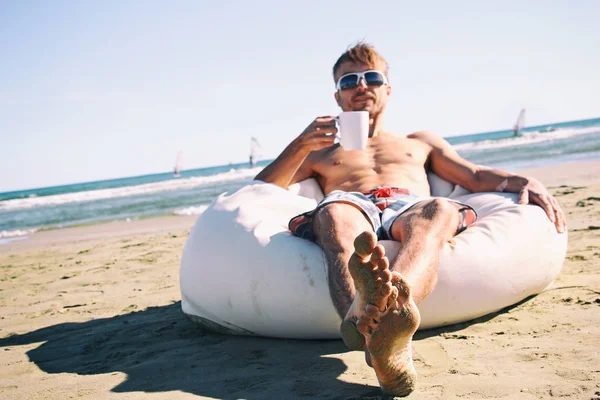 Handsome man in sunglasses and swimming pants resting on pouf on