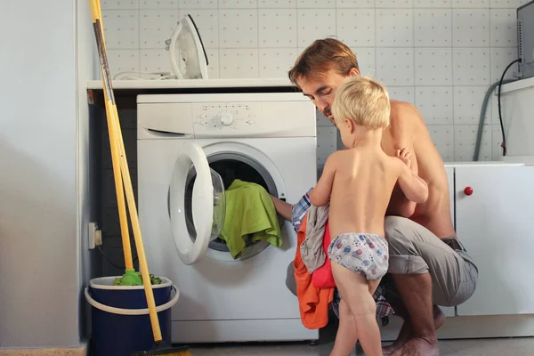 O pai e o filho mais novo fazem trabalhos domésticos. Puseram a lavandaria na máquina de lavar roupa. Trabalho em equipe familiar, conceito de dona de casa masculina — Fotografia de Stock