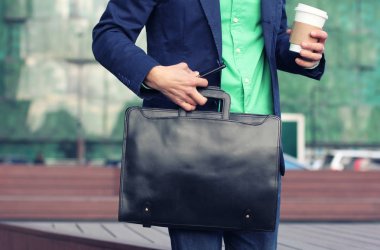 Gündelik giyim kırpılmış görüntü işadamı gitmek için kahve fincan tutmak için çalışır, deri evrak çantası ve akıllı telefon modern ofis binası arka plan karşı aynı anda.