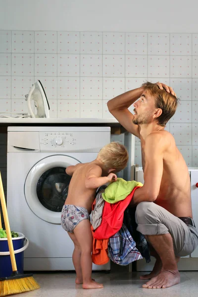 Jovem marido exausto pai sentado perto da máquina de lavar roupa com pilha de roupas em completo desespero, porque seu filho interfere em fazer tarefas domésticas. Doméstica masculina ou conceito de pai solteiro — Fotografia de Stock