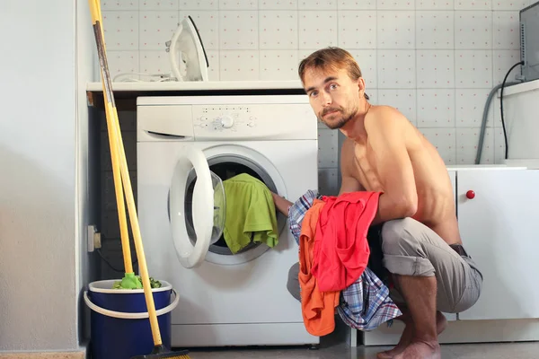 Cansado jovem caucasiano carrega as roupas sujas na máquina de lavar roupa. Trabalho doméstico de solteiro, conceito de dona de casa masculina. Homem na lavandaria . — Fotografia de Stock