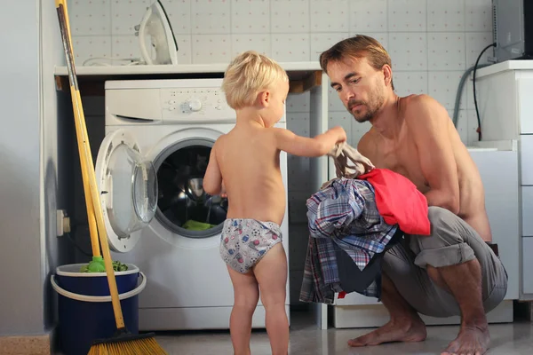 Bonito menino loiro criança ajuda seu pai a colocar a lavanderia na máquina de lavar roupa. Trabalho doméstico em família, conceito de dona de casa masculina . — Fotografia de Stock