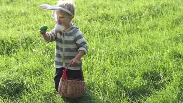 Easter egg hunting. Cute toddler caucasian boy with bunny ears finds an egg in the grass on lawn and shows it to the camera. Slow motion.
