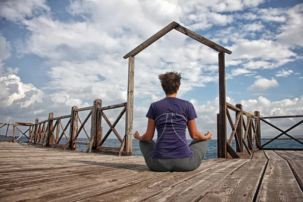 Gate to the sea. Athletic woman with short hair sitting in lotus pose on wooden pier and meditating against beautiful sky with clouds. View from behind. — Stock Photo, Image