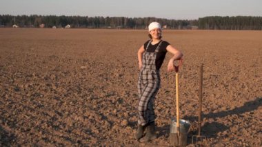 Gün batımında taze sürülmüş alanda kürek, yaba ve kova ile kadın çiftçi tam uzunlukta portre duruyor.