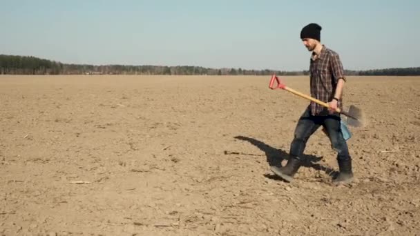 年轻的白种人留着胡子 拿着铲子和桶 走过刚刚犁过的田地 准备进行春季耕作工作 — 图库视频影像