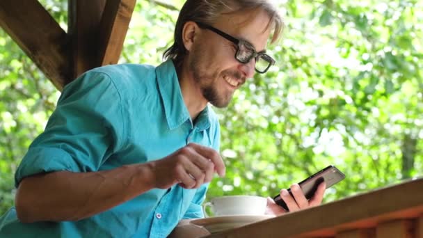英俊的胡须男子在休闲装与眼镜喝咖啡 并使用智能手机在夏季开放露台咖啡厅 — 图库视频影像