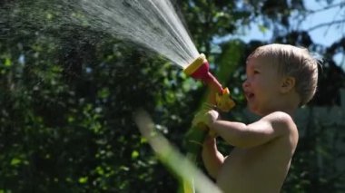 Sevimli yürümeye başlayan sarışın çocuk yaz sıcak gününde bahçe fıskiye ile oynarken. Arka bahçede eğlenen çocuk kendini hortumla püskürtüyor. Mutlu çocukluk. 