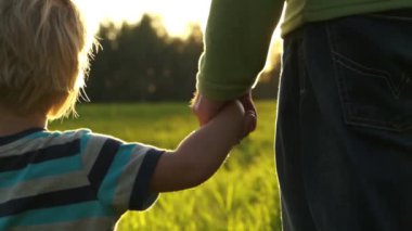 Gün batımında el ele tutuşan baba ve küçük çocuğu kapatın. Güneş ışınları parmaklarında parlar. Mutlu aile, sevgi, birliktelik.