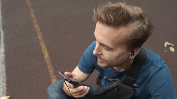 都会的なライフスタイル 都市のスポーツの遊び場に座っている間 友人とスマートフォンで話すヘッドフォンを着用した若者のクローズアップ肖像画 — ストック動画