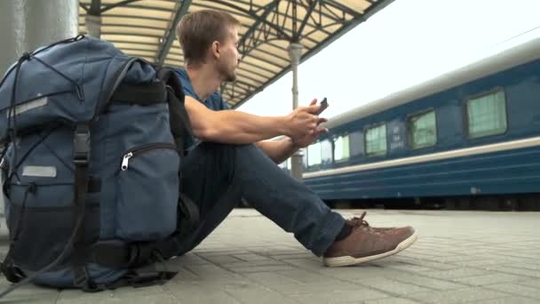 駅のホームに座って電車を待っているスマートフォンとバックパックを持つ若いあごひげの男性観光客 旅行コンセプト — ストック動画