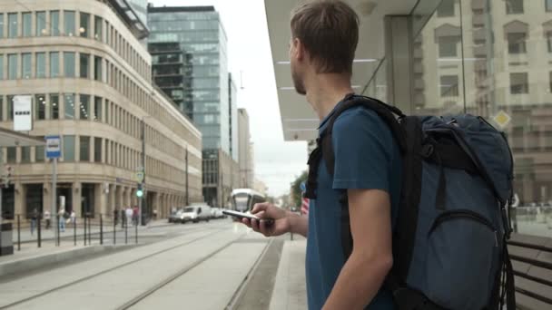 Millennial man tourist with backpack and smartphone stand on public transport stop and waiting tram in modern city center. Travel concept. — Stock Footage Millennial Man Tourist Backpack Smartphone Stand Public Transport Stop Waiting Stock Footage