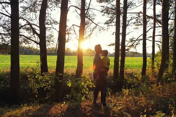 Ο πατέρας κρατά το νήπιο γιο του στην αγκαλιά του και τον φιλάει κατά του ηλιοβασιλέματος στην άκρη του δάσους. Ημέρα του πατέρα, ευτυχισμένη πατρότητα, αγγίζοντας τις οικογενειακές στιγμές. — Φωτογραφία Αρχείου