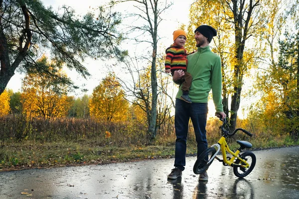 Padre y su hijo pequeño caminando en el parque en el buen día de otoño. Papá sosteniendo al niño en una mano y la bicicleta del niño en otra . — Foto de Stock