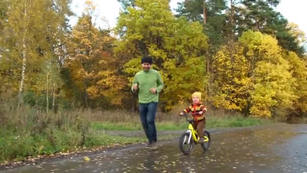 爸爸和儿子在秋天的公园玩得很开心 可爱的蹒跚学步的男孩在练习骑自行车 他的爸爸在他身边跑来跑去 给他撑腰 快乐的家庭时刻 糖果生活方式镜头 — 图库视频影像