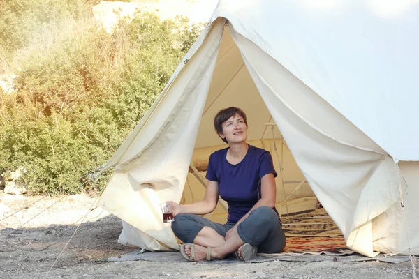 Staycations - local camping outdoor vacation. Woman drinking tea near big glamping tent with cozy interior. Luxury travel accomodation into the forest. Hyper-local travel, night camping out concept.