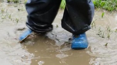 Mavi lastik botlu çocuğun ayakları oyun oynuyor ve çamur birikintisine atlıyor, yavaş çekimde kirli su sıçrıyor. Çocuk yağmurdan sonra doğada eğleniyor. Bahar ya da sonbahar mevsimsel konsepti.