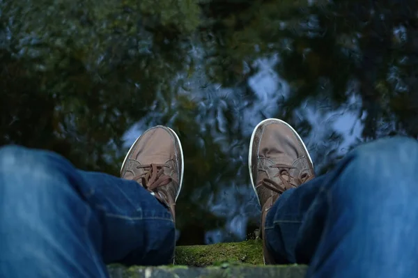 POV men feet in sneakers hanging from bridge over river. Top view male legs dangling over water. Guy enjoying time alone sitting on old bridge in park. Personal perspective, relaxing and mindfulness.