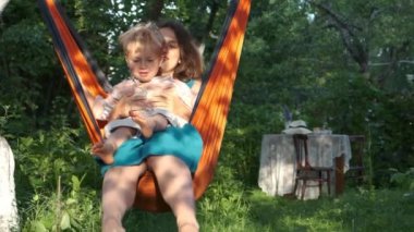 Anne ve oğlu gün batımında turuncu hamak sandalyesinde dinlenirken arka planda bahçe masası. Anne ve çocuğu yaz tatilinde köydeler. Cottagecore, kırsal kesimde yavaş yaşam. Yavaş çekim.