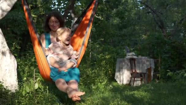母亲和儿子在日落时坐在橙色的吊床椅子上休息 背靠花园的餐桌 妈妈和孩子在村里度暑假 家庭杂务 农村生活缓慢 放松户外的观念 — 图库视频影像