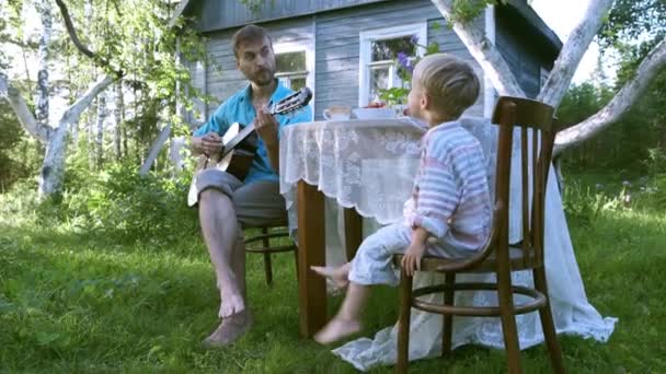 农村生活缓慢 父亲和儿子在乡间别墅附近的花园里休息 爸爸弹吉他 小孩听音乐 村里的暑假 小巧玲珑 心胸狭窄 享受小事 — 图库视频影像