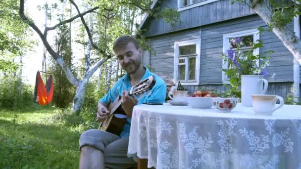 农村生活缓慢 英俊的男人在乡间别墅附近的花园里休息 坐在桌旁弹奏吉他 村里的暑假 乡下人 乡下人 乡下人 享受这些小事 — 图库视频影像