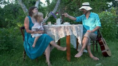 Mutlu aile babası, anne ve küçük çocuk bahçedeki masada oturuyor, böğürtlen yiyor, hasır şapkayla oynuyor. Köyde pastoral hayat, köyde yaz tatili..