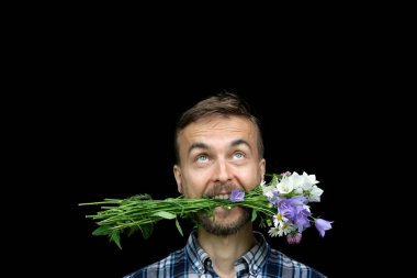 Komik sakallı, stil sahibi bir adam ağzında bir buket yabani çiçekle siyah arka plana bakıyor. Kopya alanı olan tebrik kartı, sevdiğinize hediye, bahar ya da yaz buluşması konsepti.