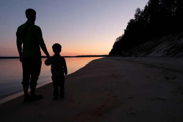 Silhouette of father and son holding hands, standing on the beach at sunset. Family on vacation. Calm water, tranquil weather, relax and meditation in evening.