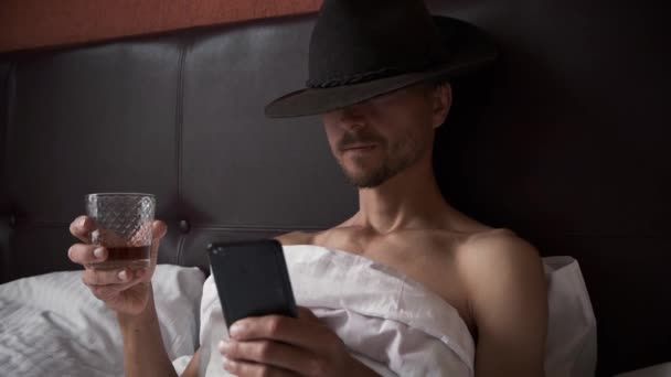 Brutalny Nieogolony Mężczyzna Kowbojskim Kapeluszu Fedory Leży Łóżku Pije Whisky — Wideo stockowe