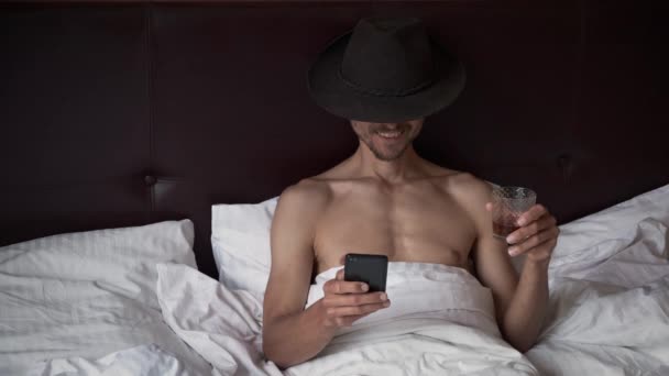 Dziwny Nieogolony Mężczyzna Kowbojskim Kapeluszu Fedory Leży Łóżku Pije Whisky — Wideo stockowe