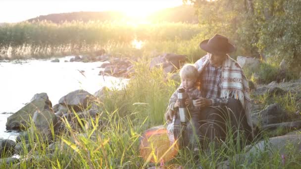 当地游客的父亲和儿子在日落时坐在湖边 用热水瓶喝热茶 与家人共度大自然时光 远足或露营生活方式 — 图库视频影像