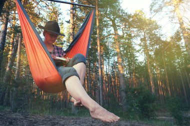 Fötr şapkalı bir kadın gün batımında hamakta otururken kitap okuyor. Farkındalık, teknoloji kırılması, refah, bilinçli yaşam, kişisel büyüme konsepti.