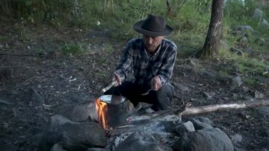 Yaz ormanlarında kamp ateşinde yemek pişiren yakışıklı bir turist. Keçeli şapkalı hippi sakallı adam ateşte demlikte su kaynatıyor. Ormanda kamp ve vahşi maceralar..