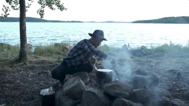 游客徒步旅行者在湖边的营地生火做饭 希斯特留着胡子的人戴着毛毡帽 在火上把水放在锅里烧开了 秋天的野营 森林中的野生冒险 — 图库视频影像